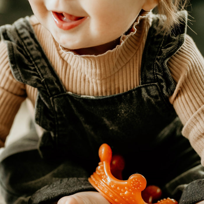 A young child wearing a beige ribbed sweater and dark overalls smiles while holding the Haakaa 100% Food Grade Silicone Sensory Development Crown Teether in orange. The focus is on the childs joyful expression, capturing a playful moment that encourages sensory development.