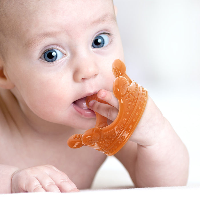 A baby with bright blue eyes lies down, chewing on a Haakaa 100% Food Grade Silicone Sensory Development Crown Teether. The soft, out-of-focus background highlights both the baby and the colorful teething toy.