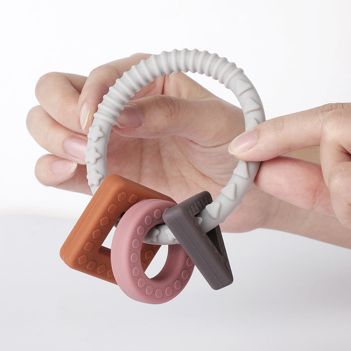 Hands holding Haakaas 100% Food Grade Silicone Sensory Development Teething Ring, featuring three BPA-free textured charms for teething relief: a rust-colored square, a pink circle, and a gray triangle. The gray ribbed ring combines style and functionality.