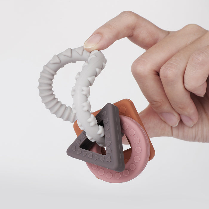 A hand holds the Haakaa 100% Food Grade Silicone Sensory Development Teething Ring, featuring BPA-free silicone with multiple rings and shapes, including triangular and circular elements in gray, pink, and orange for gentle teething relief with textured surfaces for sensory stimulation.