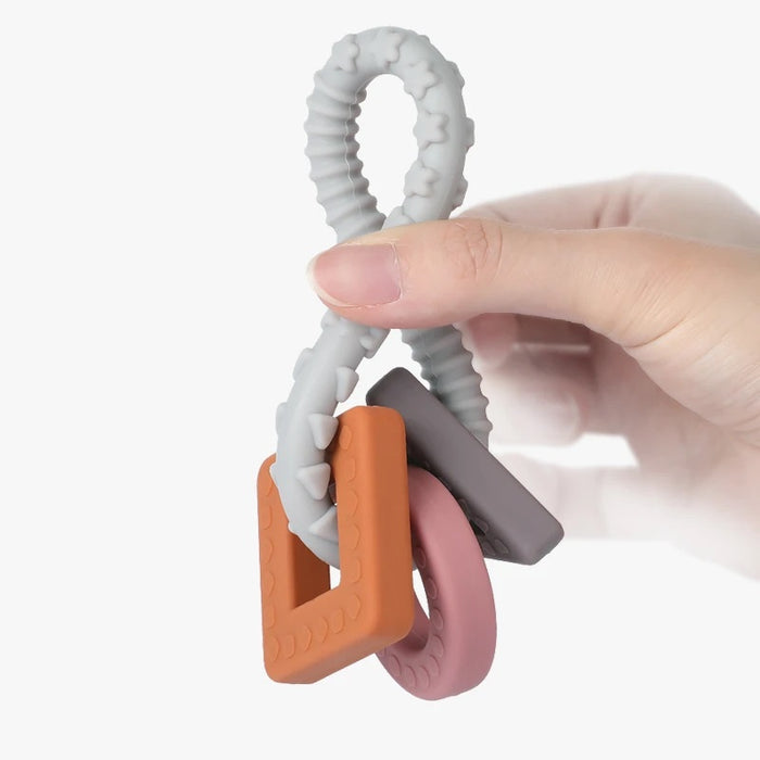 A hand holds the Haakaa 100% Food Grade Silicone Sensory Development Teething Ring, featuring three textured rings in gray, orange, and pink on a looped handle, providing soothing relief for little ones against a crisp white backdrop.