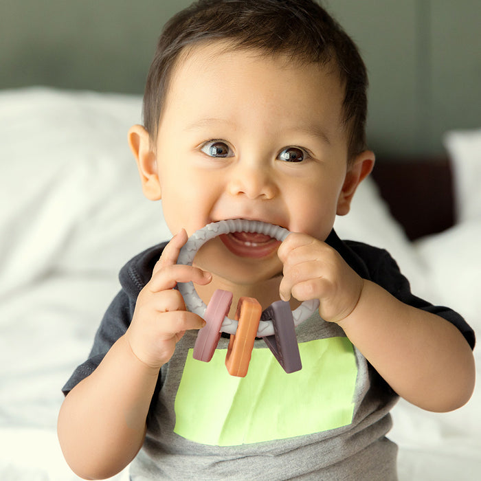 A baby with short dark hair smiles while biting a Haakaa 100% Food Grade Silicone Sensory Development Teething Ring, featuring three colorful rectangular blocks for soothing relief. Wearing a gray shirt with a green square, the baby is set against a softly blurred background.