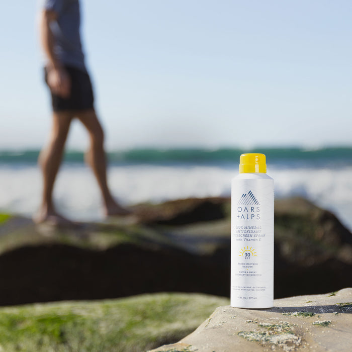 A bottle of Oars + Alps 100% Mineral Antioxidant Sunscreen Spray - SPF 30 rests on a rock by the ocean under bright sunlight, as a blurred figure in shorts walks nearby, emphasizing its antioxidant-rich formula.