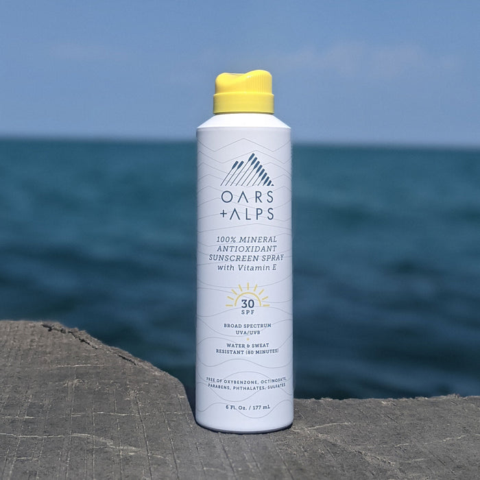 A can of Oars + Alps 100% Mineral Antioxidant Sunscreen Spray - SPF 30 with a yellow cap sits on wood, ocean and blue sky behind. The label highlights mineral sunscreen protection by Oars + Alps.