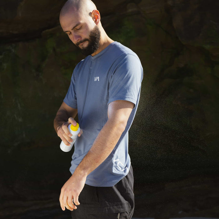 A bearded, shaved-head man in a light blue T-shirt and black pants applies Oars + Alps 100% Mineral Antioxidant Sunscreen Spray - SPF 30 to his forearm outdoors against a dark rocky background.