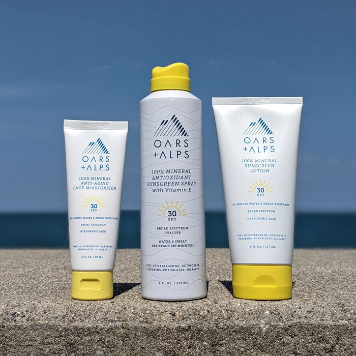 Three Oars + Alps 100% Mineral Antioxidant Sunscreen Sprays with yellow caps and SPF 30 are displayed on a stone ledge outdoors, set against a blue sky and lake backdrop.
