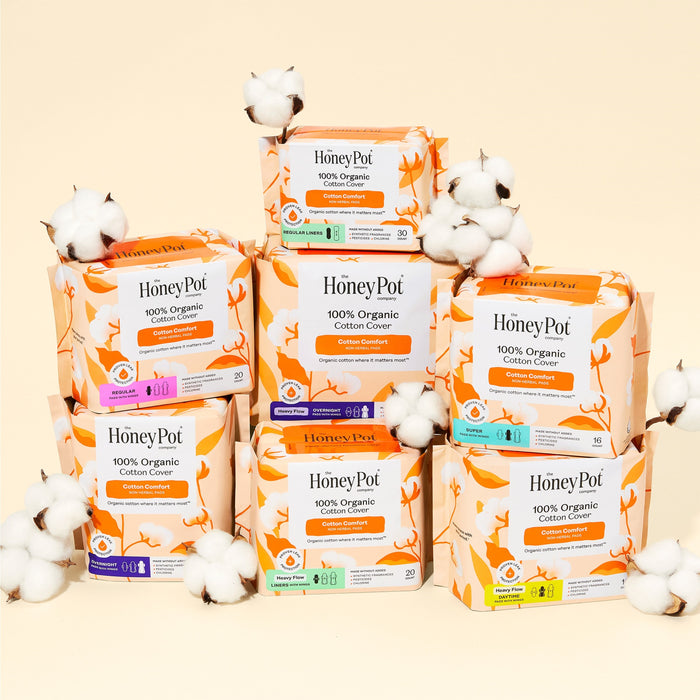 Seven packs of The Honeypot Company 100% Organic Cotton Cover Pads are stacked together among cotton flowers on a beige background. These regular pads with wings deliver gentle, reliable menstrual protection.
