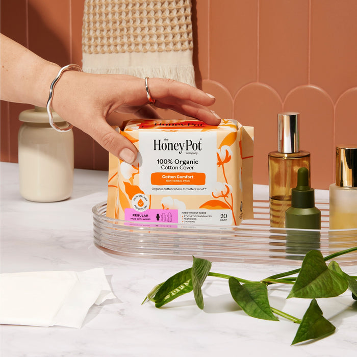A hand reaches for a box of The Honeypot Company 100% Organic Cotton Cover Pads on a tray with skincare bottles, a plant, and a packet on white marble—a perfect choice of regular pads with wings for everyday comfort.