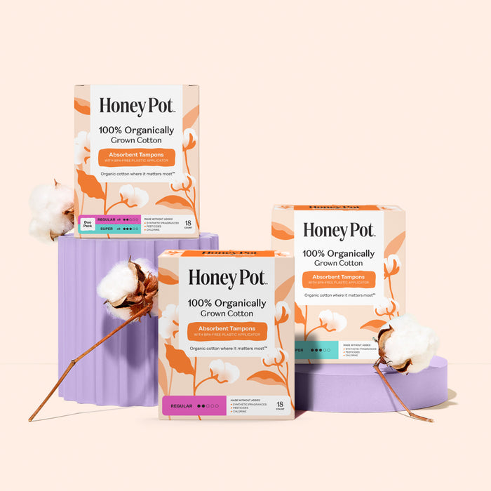 Three boxes of The Honeypot Company 100% Organic Regular Tampons are displayed on purple stands with cotton branches, against a light peach background, highlighting their trusted leak protection.