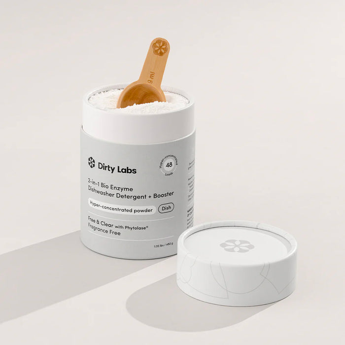 An open container of Dirty Labs 2-in-1 Bio Enzyme Dishwasher Detergent + Booster with a wooden scoop inside, placed beside its lid on a neutral background. This bioenzyme-powered formula delivers powerful cleaning and a spotless finish in every wash.