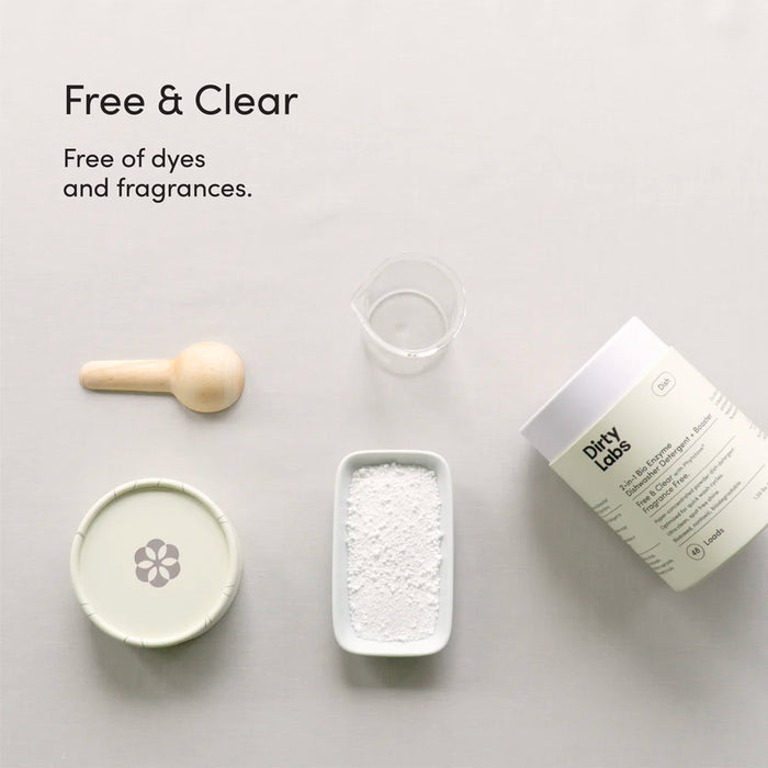A container of Dirty Labs 2-in-1 Bio Enzyme Dishwasher Detergent + Booster, a wooden scoop, a small beaker, and a dish of white powder are arranged on a light surface. Text reads: “Free & Clear. Free of dyes and fragrances.” Powered by bioenzymes and our clean rinse formula for effective results.