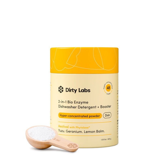 A yellow container of Dirty Labs 2-in-1 Bio Enzyme Dishwasher Detergent + Booster sits beside a wooden spoon filled with white powder, all set against a white background.
