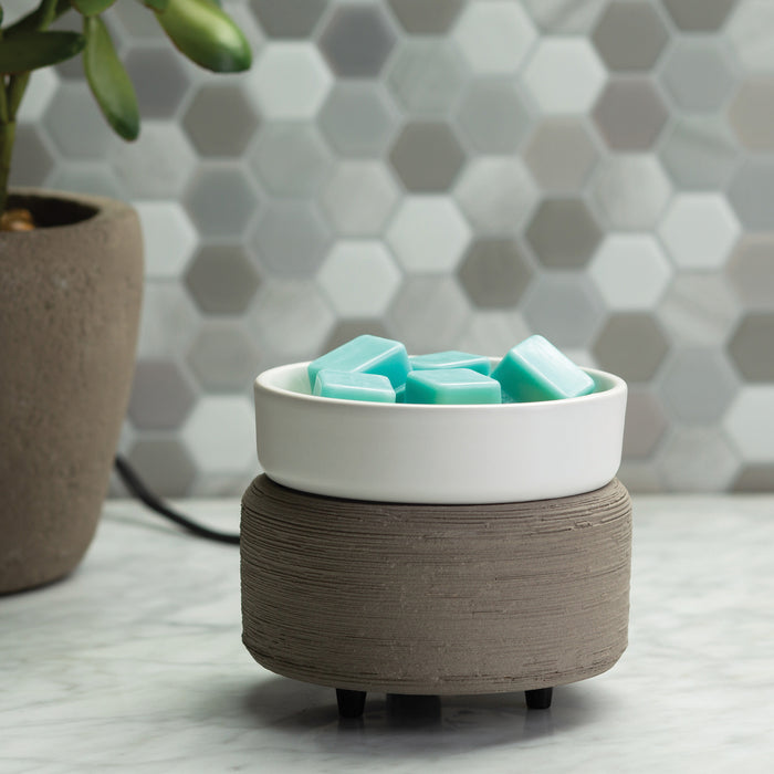 On a marble surface, Candle Warmers Etc.s 2-in-1 Classic Warmer in gray ceramic with a white dish holds turquoise wax melts. A hexagonal tile wall and potted plant add charm, while this versatile warmer doubles as a stylish companion for jar candles to create an inviting ambiance.