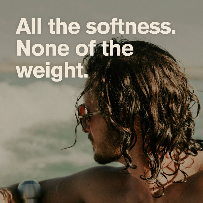A person with wet, wavy hair and sunglasses sits outdoors by water. Bold text reads, All the softness. None of the weight. Perfect for men’s grooming, this look is easy to achieve with Every Man Jack 2-in-1 Shampoo and Conditioner for Men, Naturally Derived—featuring an invigorating tea tree formula.