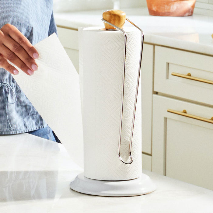Wearing a denim shirt, someone pulls a sheet from Grove Co.s Bamboo Paper Towels (140 sheets per roll) on a stand. The stand sits on a white kitchen countertop with light-colored cabinets in the background, emphasizing sustainable living.