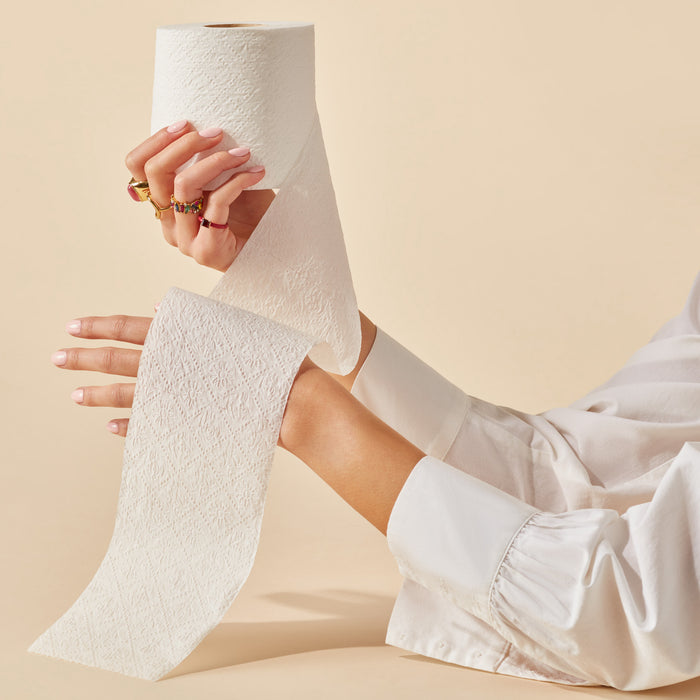 A person with multiple rings holds Caboos 2-Ply Bath Tissue against a beige background. Partially unrolled, the bamboo, tree-free toilet paper is presented by someone in a white shirt.