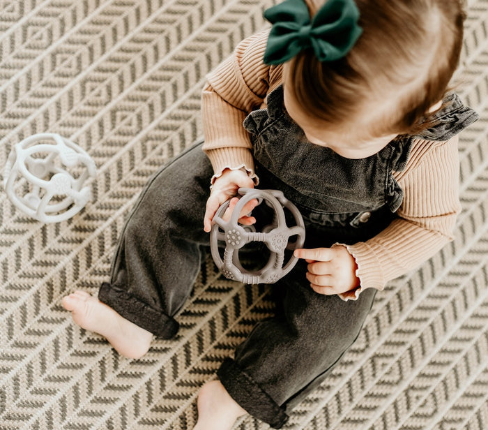 A toddler with a green bow in their hair sits on a patterned rug, playing with a BPA-free gray toy. They wear a striped shirt and dark overalls, surrounded by Haakaas FDA-approved 100% food grade silicone teething ball.