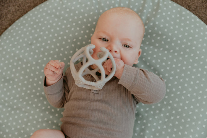 A baby lies on a green mat with white stars, wearing a brown outfit and holding the Haakaa 100% Food Grade Silicone Teething Ball. The child gazes curiously at the gray, BPA-free toy.