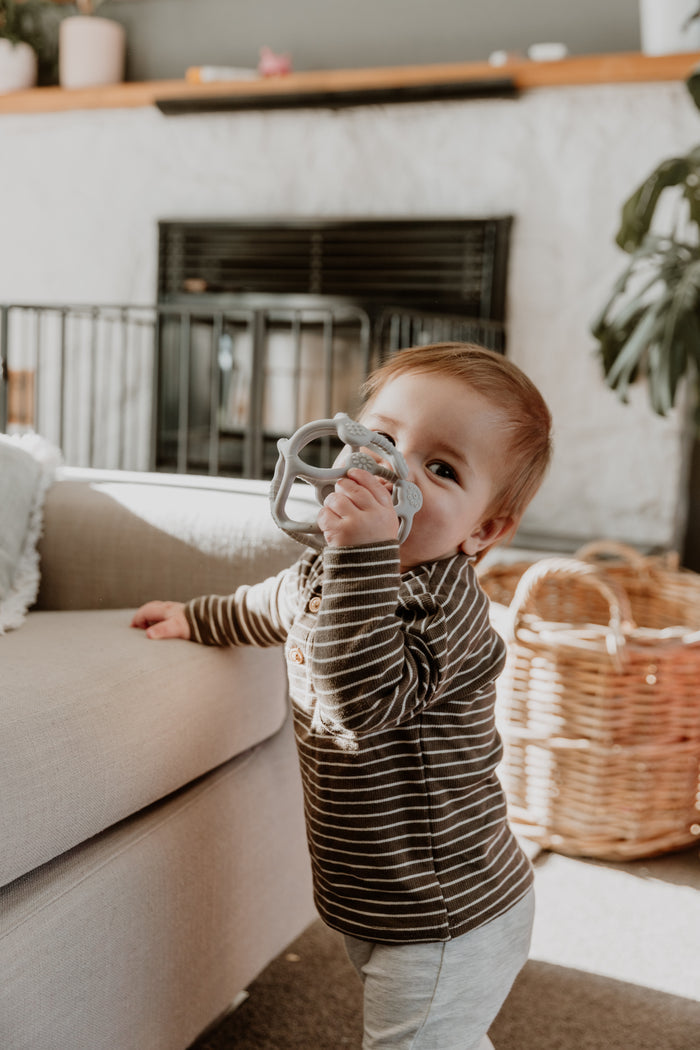 A toddler in a brown-striped shirt stands next to a beige couch, holding an FDA-approved Haakaa 100% Food Grade Silicone Teething Ball near their mouth. A wicker basket is nearby, with a plant and fireplace behind them, creating a cozy living room scene.