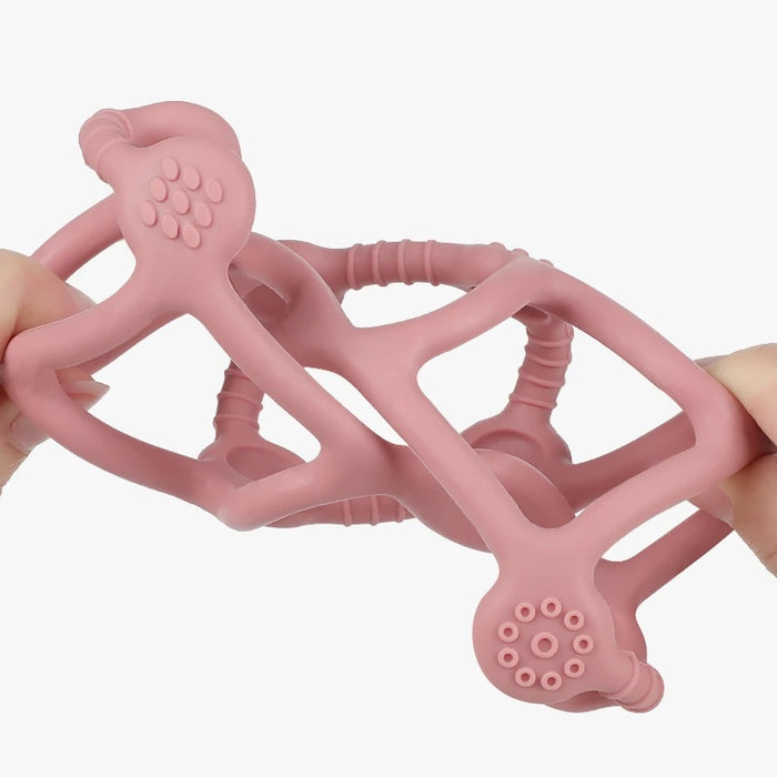 Two hands stretch a pink, flexible Haakaa 100% Food Grade Silicone Teething Ball with honeycomb-like ends and a geometric lattice design. BPA-free and FDA-approved, its set against a pristine white background.