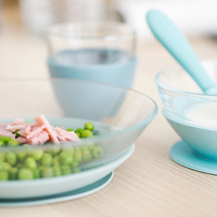On a light wooden table, BEABAs eco-friendly 3-Piece Glass Suction Baby and Toddler Feeding Set holds green peas with ham in one bowl and yogurt in another with a blue spoon. A pastel blue drinking glass is in the background.