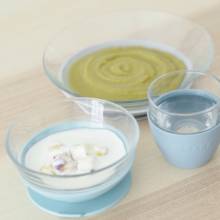 The eco-friendly tableware set includes BEABAs 3-Piece Glass Suction Baby and Toddler Feeding Set: a clear bowl with yogurt and fruit, another bowl of green puree, and a Duralex glass of water in a light blue holder, all on a light wood table.