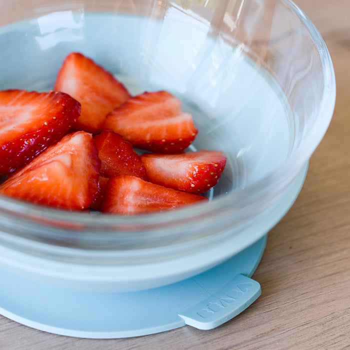 Sliced strawberries are in a clear BEABA Duralex bowl with a blue silicone base on wood. This eco-friendly 3-Piece Glass Suction Baby and Toddler Feeding Set has BMMA embossed on the side, enhancing any kitchen while promoting sustainability.