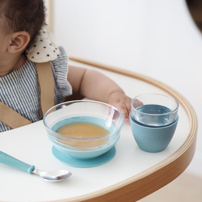 A baby in a striped outfit sits eagerly in a high chair, surrounded by BEABAs 3-Piece Glass Suction Baby and Toddler Feeding Set. A bowl with food, spoon, and cup with a blue cover rest on the tray as sunlight brightens the minimal setting.