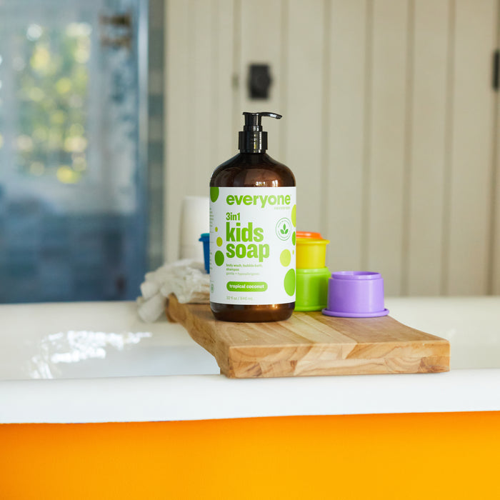 A bottle of 3in1 Kids Soap Shampoo & Bubble Bath by Everyone, infused with essential oils and a tropical coconut scent, sits on a wooden tray by the bathtub. Nearby, colorful stacking cups brighten the space as natural light fills the bathrooms crisp white walls.