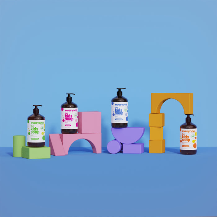 Four bottles of Everyones 3in1 Kids Soap Shampoo & Bubble Bath rest on vibrant geometric foam shapes against a blue backdrop. Each bottle features a unique scent, like lavender lullaby and orange squeeze, and the colorful shapes are green, pink, purple, and yellow.