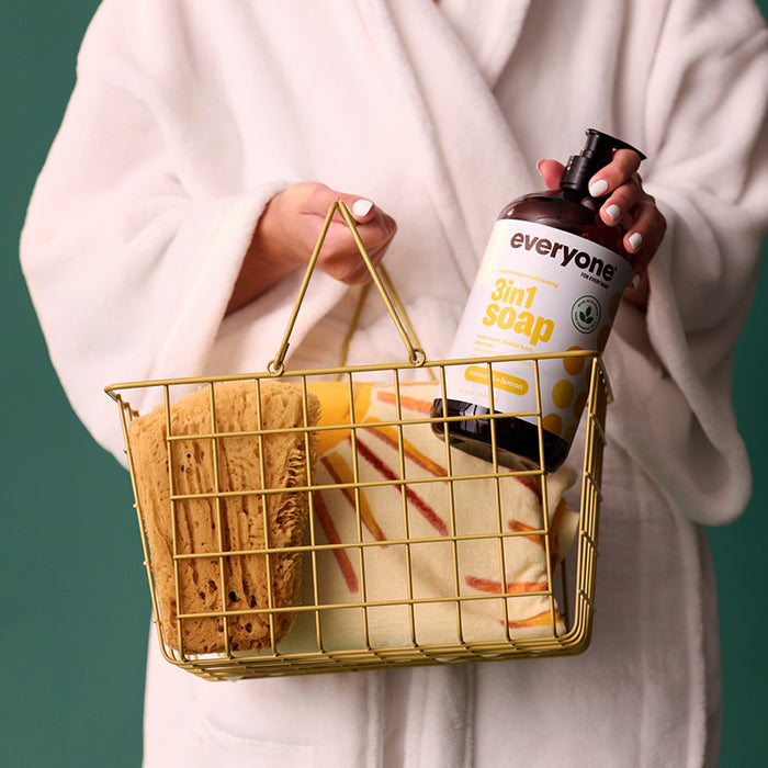 A person in a white robe holds a gold wire basket with sponges and Everyone 3in1 Soap, Shampoo, & Bubble Bath infused with essential oil. The soothing green background enhances the calmness and care atmosphere.