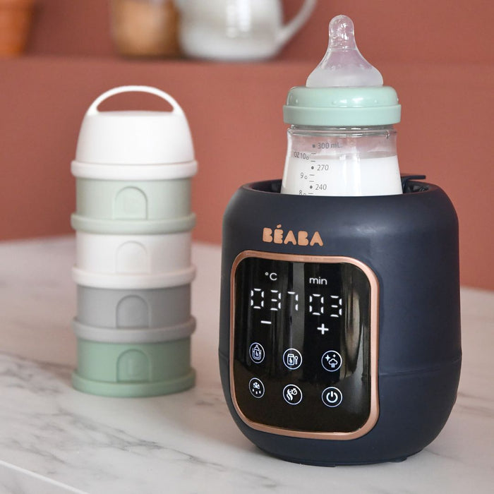 The BEABA 5-in-1 Multi Milk Baby Bottle and Food Warmer with a digital display holds baby bottles. Nearby, a stackable container with four compartments in white, grey, light grey, and mint green sits on a marble countertop next to baby food jars.