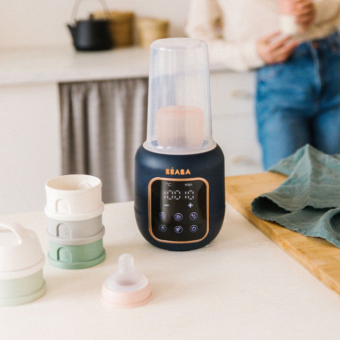 On the kitchen counter, a BEABA 5-in-1 Multi Milk Baby Bottle and Food Warmer with LED display stands poised to heat baby’s bottles. Nearby, silicone cup lids and a baby bottle nipple are ready for use as someone in jeans and a light sweater leans against the counter.