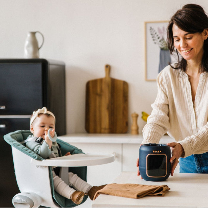 A woman smiles while using a BEABA 5-in-1 Multi Milk Baby Bottle and Food Warmer on the counter. A baby in a high chair nearby holds a spoon, with a black fridge and decorative wall items in the background.