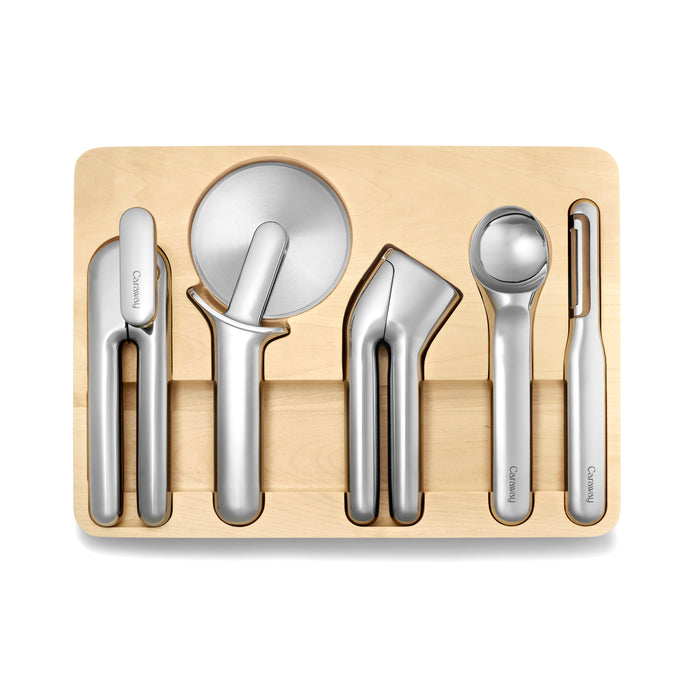 The Caraway 5-Piece Stainless Steel Kitchen Gadget Set includes a peeler, pizza cutter, garlic press, ice cream scoop, and can opener. Each tool has an ergonomic design and fits neatly into its designated slot on the wooden tray.