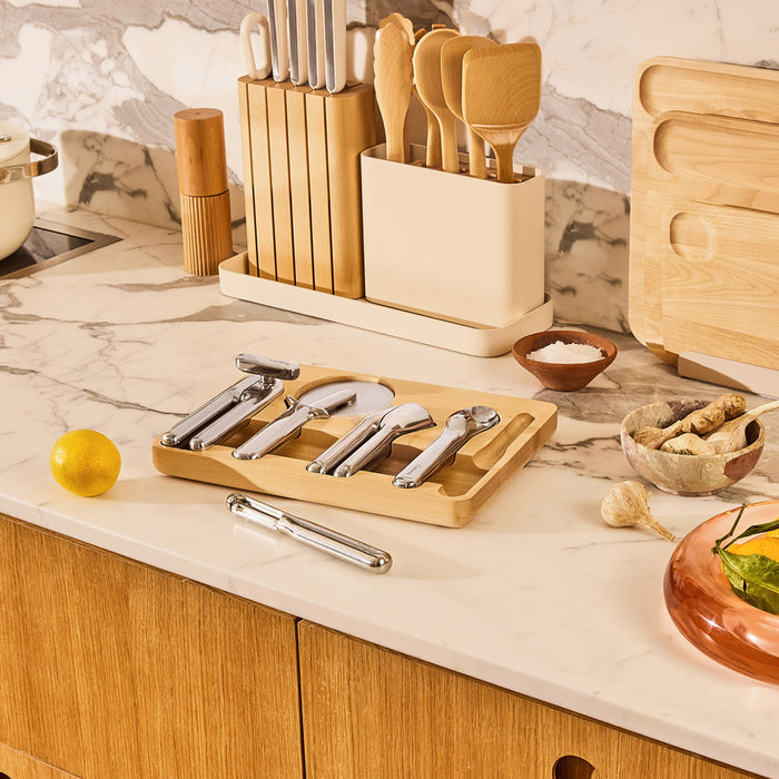 A marble kitchen counter displays a knife set, lemon, ginger, garlic, and a bowl of salt, accompanied by Caraways 5-Piece Stainless Steel Kitchen Gadget Set. Wooden cabinets are below and mixed among the essentials is an ergonomic kitchen tool in the foreground.