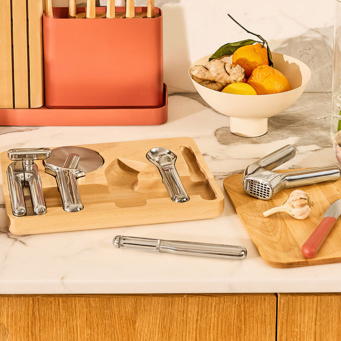 A kitchen countertop features a wooden tray displaying the Caraway 5-Piece Stainless Steel Kitchen Gadget Set, including a peeler and garlic press. Nearby, a bowl of oranges and ginger sits with a red knife and garlic clove, emphasizing the elegance of essential kitchen tools.