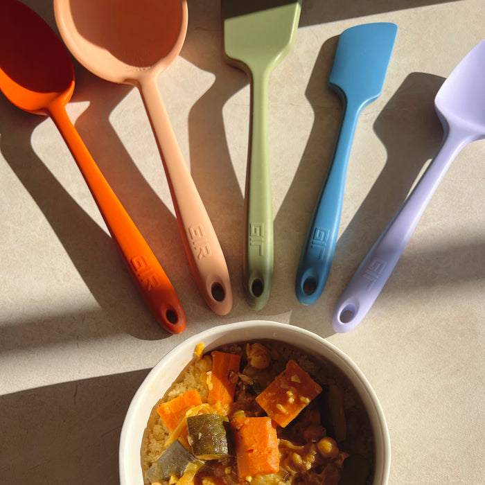 On a light countertop, a beige bowl filled with hearty vegetable stew sits beneath five colorful spatulas from the GIR 5-Piece Ultimate Tool Set, arranged in a fan pattern.