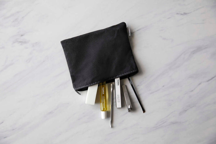 A black makeup pouch on a marble surface, partially unzipped, reveals various cosmetic items including Terra Beauty Bars yellow cuticle oil, their 7-Piece Manicure Kit in a small white container, a black pencil, and two rectangular boxes.