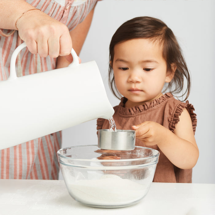 A young child holds a measuring cup as an adult pours water from a white pitcher into it over a flour-filled glass bowl, using the Elseware Unplug A Day at the Beach Set—a unique baking kit inspired by beach treasures.
