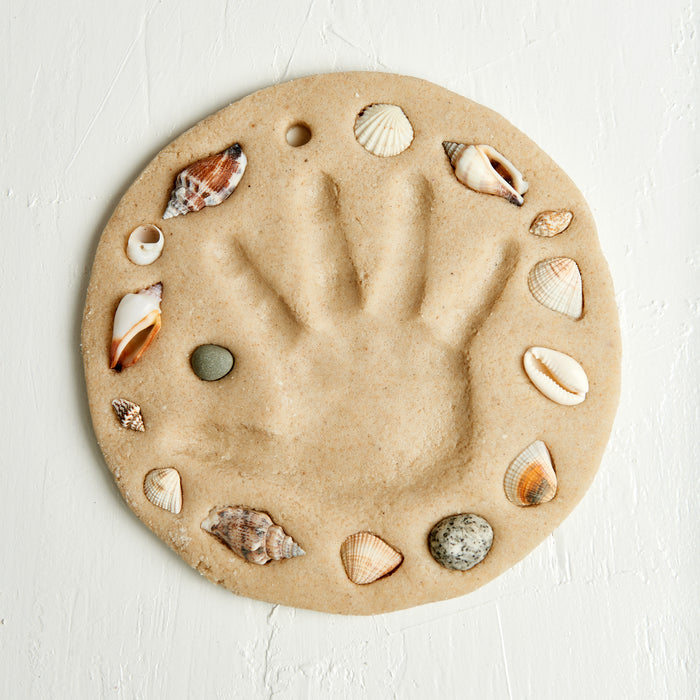 The Elseware Unplug A Day at the Beach Set features a sand-colored, round clay piece with a central handprint, edged with seashells and a small stone—perfect for capturing beach memories.