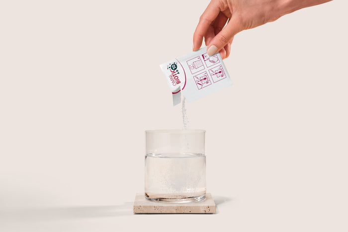 A hand expertly pours Omni-Biotics AB10 powder from a small packet, promising enhanced bacterial diversity, into water on a stone coaster. Illustrated instructions decorate the probiotic supplement packet against a beige backdrop.