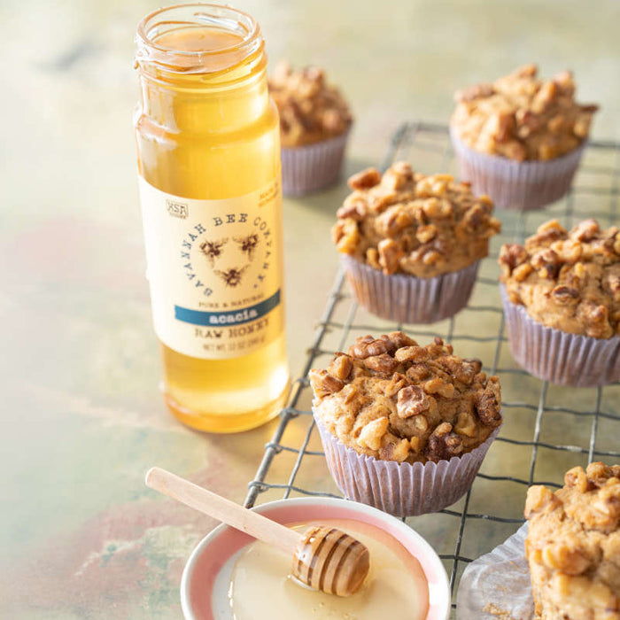 A jar of Savannah Bee Companys Acacia Honey sits next to nut-topped muffins on a cooling rack. A wooden dipper on a plate with Raw Acacia Honey, renowned for its high fructose content, adds sweetness in the foreground.
