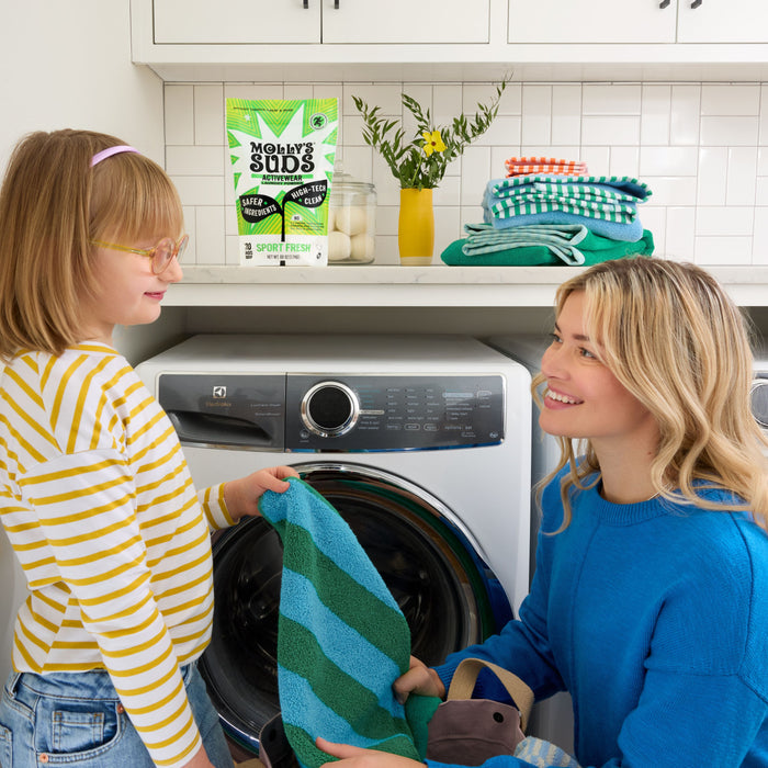 A woman and a girl smile as they do laundry together near a front-loading washer with colorful towels. Molly's Suds Activewear Laundry Powder for odor removal and neatly folded towels are displayed on the counter behind them.