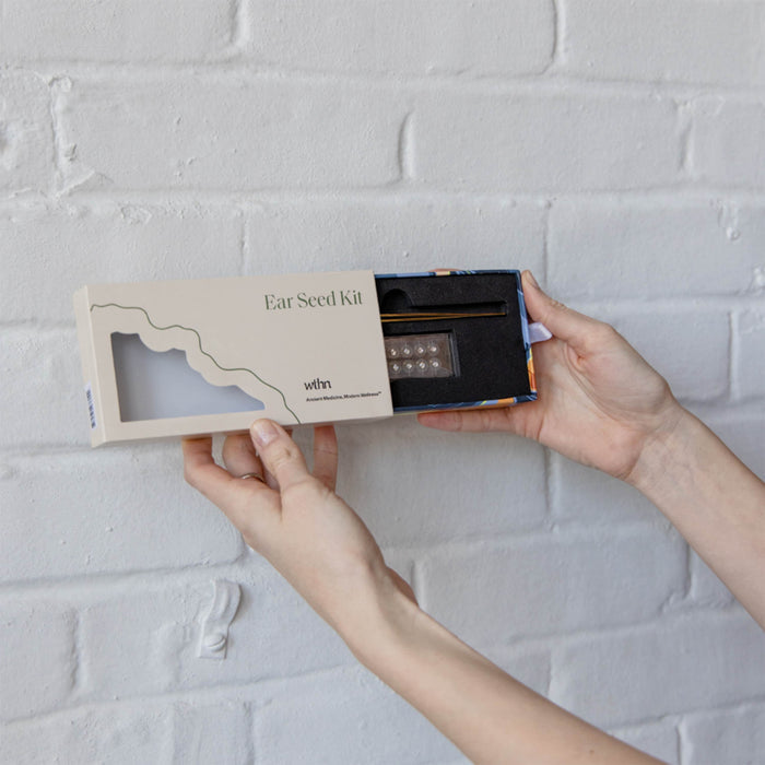 Hands hold an open Acupressure Crystal Ear Seed Kit against a white brick wall. The packaging shows Acupressure Crystal Ear Seed Kit and WTHN. Inside are ear seeds and a placement tool for acupressure-based pain relief.