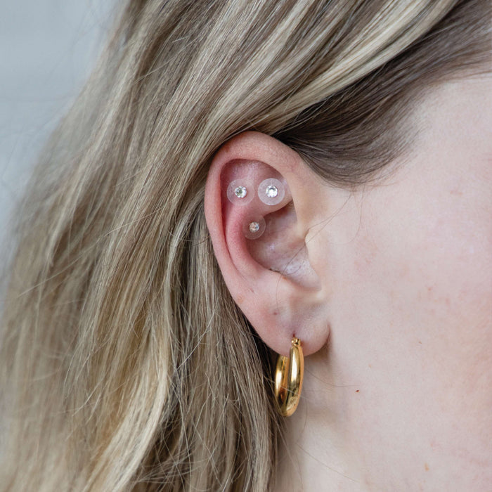 A close-up shows a persons ear with WTHN silver acupressure crystal ear seeds in the cartilage and a gold hoop in the lobe. The light blond hair frames the ear, offering style and potential pain relief similar to ear seed therapy.