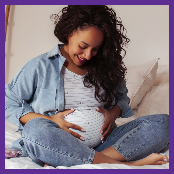 A pregnant woman sits on a bed, smiling and gently cradling her baby bump, promoting healthy fetal development. She wears a striped shirt and denim overalls in a cozy space with New Chapters Advanced Perfect Prenatal™ Multivitamin for Healthy Baby & Mom nearby, framed by a soft purple border.