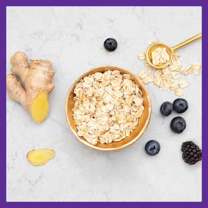 A wooden bowl of oats sits on marble, surrounded by ginger, blueberries, and a blackberry. A gold spoon glimmers nearby. The scene in purple tones echoes the vibrant support of New Chapters Advanced Perfect Prenatal™ Multivitamin for Healthy Baby & Mom.