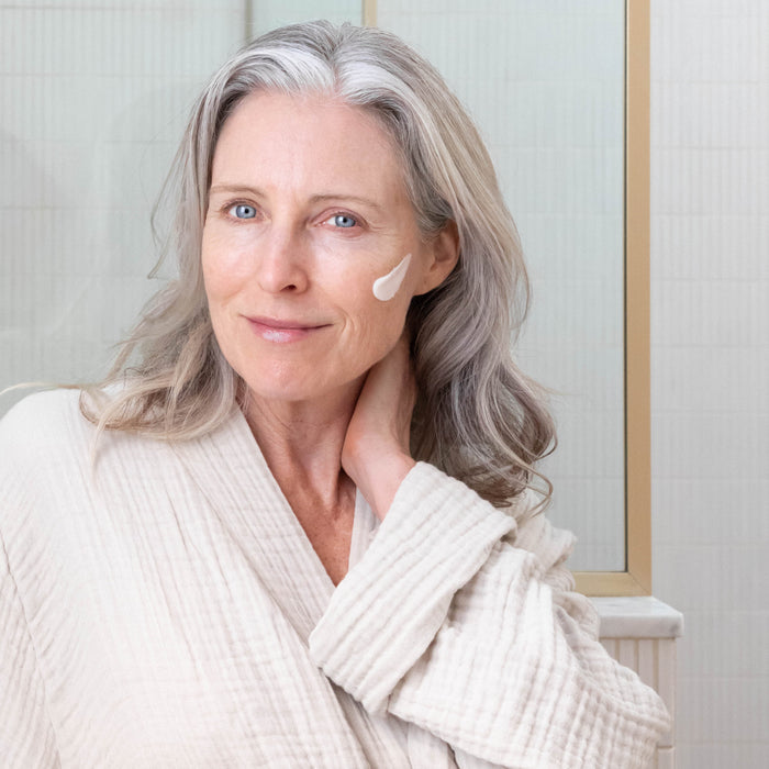 A smiling older woman with long gray hair wears a cream-colored robe and applies OSEA’s Advanced Protection Cream to her cheek while standing in a bright bathroom.