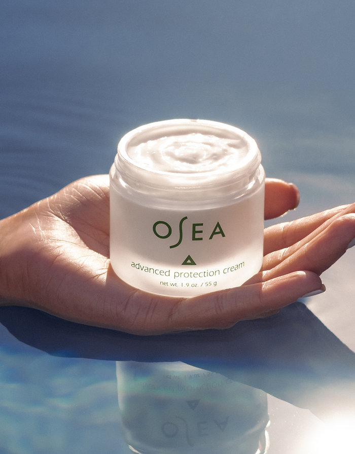 A hand holds an open jar of OSEA Advanced Repair Eye Cream above a blue water surface, with sunlight glistening off the water to create a serene atmosphere that highlights its benefits in reducing puffiness.